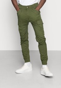 Olivgrüne Cargo-Hosen mit elastischen Bündchen und mehreren Taschen, gefertigt aus einem glatten Stoff, kombiniert mit weißen Sneakers und sichtbaren Bündchen.