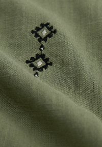 Tessuto verde con ricamo a forma di diamante in bianco e nero. La texture è leggermente ruvida con un motivo intrecciato.