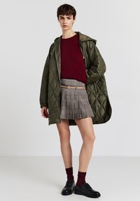 Manteau matelassé vert olive porté sur un pull bordeaux, associé à une jupe plissée à carreaux et des chaussures noires. L'ensemble comprend une ceinture et des chaussettes rouge foncé.