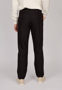 Pantalon noir à coupe ajustée, avec deux poches arrière et une texture lisse, associé à des baskets de couleur claire.
