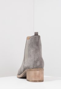 Botines de ante gris con paneles laterales elásticos, punta redondeada y tacón apilado de madera. Vista trasera que muestra una lengüeta para facilitar su colocación.