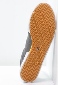 Semelle de chaussure avec motif de marche couleur beige mettant en avant le logo et le nom Tommy Hilfiger, semelle intermédiaire blanche, et dessus gris visible de profil.