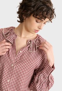 Camicia a quadri rossi e crema con colletto arricciato, chiusura a bottoni e maniche a sbuffo, realizzata in tessuto leggero.