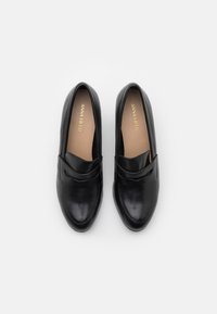 Černé kožené slip-on boty s kulatou špičkou, mají široký pás přes přední část a hnědé vložky s zlatým logem uvnitř.