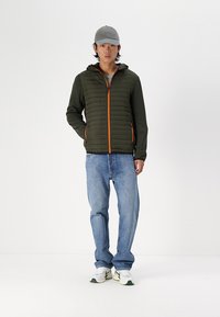 Olivgrüne Pufferjacke mit orangefarbenem Reißverschluss, getragen über einem grauen Shirt, kombiniert mit blauen Jeans und weißen Sneakern. Das Model trägt eine graue Mütze.