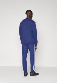 Jūras spēku sporta tērps ar baltām svītrām uz piedurknēm un biksēm, izgatavots no gludas auduma. Persona valkā melnus sporta apavus.