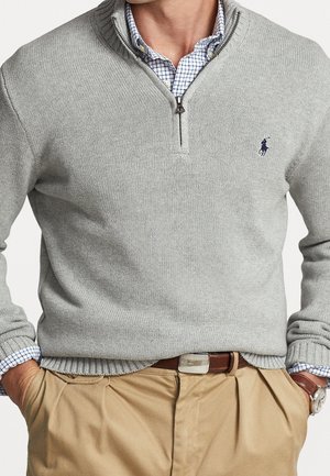 Man wearing grey quarter-zip sweater over blue checkered shirt, beige pants with brown leather belt and wristwatch, hands in pockets.