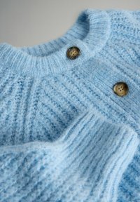 Gaiši zils adīts džemperis ar teksturētu rakstu, ar ribotām aprocēm un divām apaļām brūnām pogām apkaklē.