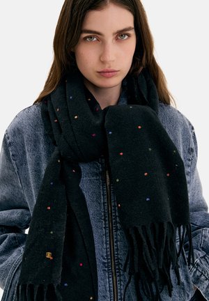 Joven mujer lleva una bufanda negra con flecos y puntos de colores sobre una chaqueta vaquera azul, mirando al frente con expresión neutra.