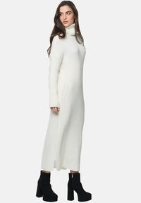 Robe en turtleneck tricot à manches longues et à rayures, blanche, avec des fentes sur les côtés. Associée à des bottes à talons épais noires.