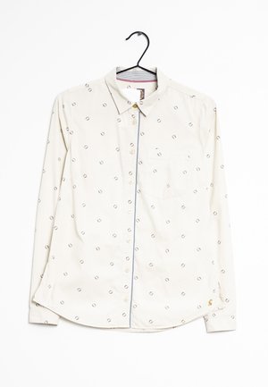 Chemise blanche à manches longues avec petits motifs circulaires, une poche poitrine avant et un col, suspendue sur un cintre noir sur un fond blanc.