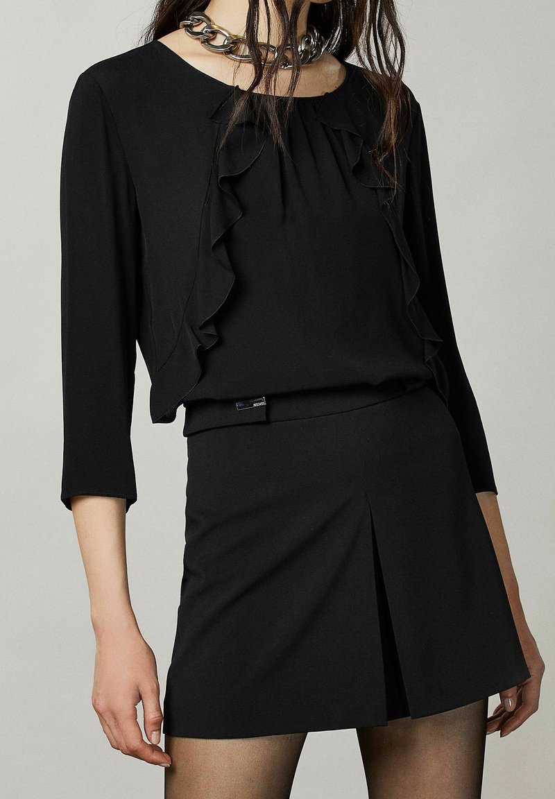 Outfit nero composto da una blusa con volants a maniche lunghe e una mini gonna aderente. Tessuto liscio, design semplice e un'etichetta ben visibile in vita.