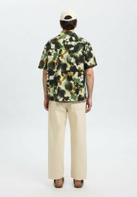 Kurzärmliges Hemd mit Knöpfen in grün, braun und schwarzem abstraktem Muster; beiger weit geschnittene Hose; Person trägt eine hellfarbene Mütze und Sandalen.