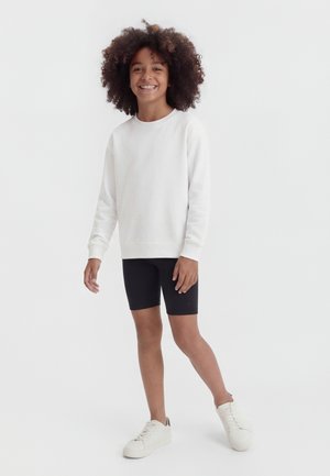 Smilende barn med krøllet hår står iført hvid langærmet skjorte, sorte shorts og hvide sneakers mod en ensfarvet baggrund.