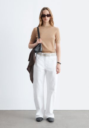 Moteris dėvi smėlio spalvos trumpomis rankovėmis palaidinę, baltas aukšto liemens kelnes, juodus batus, akinius nuo saulės, neša juodą petinę krepšį su dekoratyviniu šaliu.