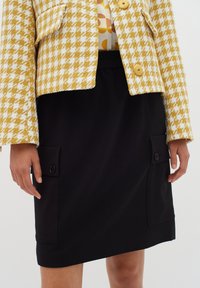 Keltainen ja valkoinen houndstooth-lyhennettävä takki suurilla nappeilla mustan hameen päällä, jossa on etupocketsit nappikoristeilla.