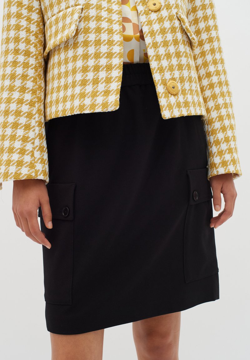 Keltainen ja valkoinen houndstooth-lyhennettävä takki suurilla nappeilla mustan hameen päällä, jossa on etupocketsit nappikoristeilla.