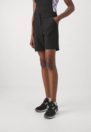 Osoba ubrana w czarne szorty z bocznymi kieszeniami, czarno-białe buty sportowe, stojąca na jednolitym jasnym tle.