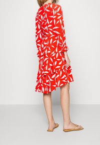 Rotes Kleid mit weißem Blattmuster, lange Ärmel, rüschendes, gestuftes Design, weicher Stoff, kombiniert mit hellbraunen flachen Sandalen, von hinten betrachtet.