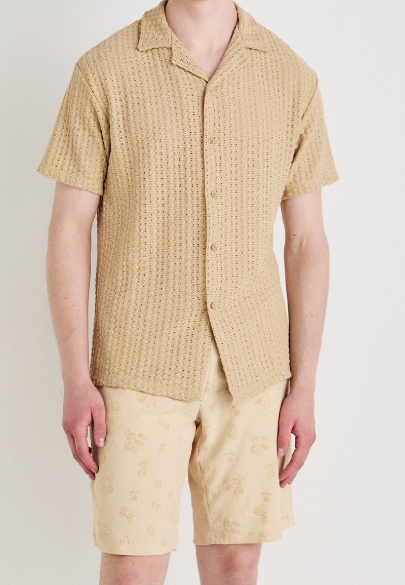 Korte mouwen, overhemd met knopen in een tan kleurige textuurstof met een subtiel patroon. Gecombineerd met beige shorts met een bloemenpatroon.