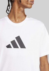 Camiseta de algodón blanca con un gran diseño de tres rayas negras en el pecho. Manga corta, forma clásica de cuello redondo.