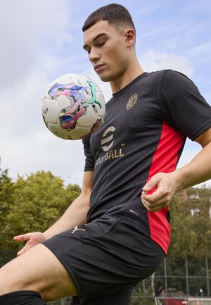 Maglia sportiva nera e rossa con logo e etichetta drycell, abbinata a pantaloncini. Un pallone da calcio a motivi è bilanciato sul ginocchio del giocatore.