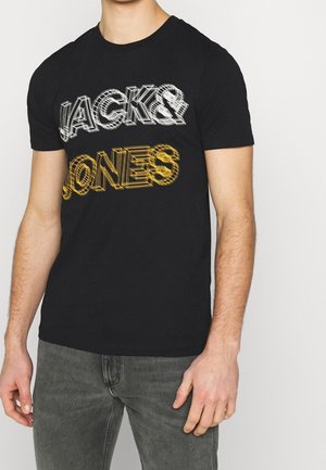 Hombre con camiseta negra que lleva texto en estilo 3D blanco y amarillo que dice "JACK & JONES", combinado con jeans grises.