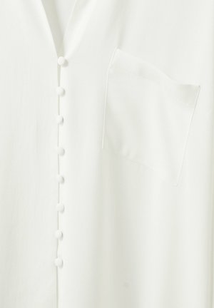 Valkoinen vaatekappale, jossa on kankaan peittämät napit etuosassa ja neliskulmainen rintatasku oikealla puolella.