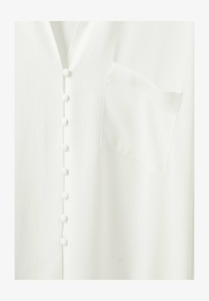 Valkoinen vaatekappale, jossa on kankaan peittämät napit etuosassa ja neliskulmainen rintatasku oikealla puolella.