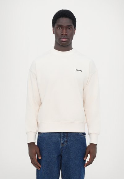 Sweatshirt couleur crème en matériau doux, avec un col rond, des poignets côtelés et un logo "BARROW" noir sur la poitrine. Jean bleu.