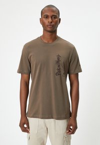 Olivenfarvet bomulds T-shirt med korte ærmer, rund halsudskæring og et grafisk print på venstre side, der viser tekst og abstrakt design.