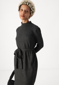 Γκρι πλέκτο φόρεμα με ψηλό λαιμό, μακριά μανίκια και ζώνη με κόμπο στη μέση. Διαθέτει απαλή υφή και χαλαρή σιλουέτα.