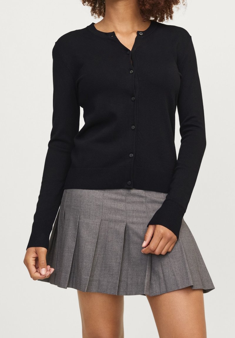 Svart stickad kofta med rund hals, knäppning, långa ärmar och ribbad nederkant, kombinerad med en grå plisserad kjol.