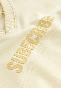 Sweatshirt couleur crème avec un tissu texturé, doté d'une inscription en lettres dorées en caoutchouc qui épelle "SUB5CRIBE" de manière proéminente sur le devant.