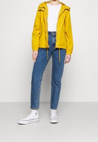 Żółta lekka kurtka z kapturem, ściągaczami i elastycznymi mankietami, w połączeniu z niebieskimi dżinsami i białymi wysokimi trampkami.