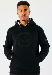 Hombre con cabello corto y rizado que lleva una sudadera negra con el logo "Project X Paris", con las manos en los bolsillos, de pie frente a un fondo claro y liso.