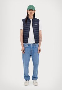 Jūras spēku puffera veste, balts krekls, gaiši zili platāki džinsi un bēša kedas ar zaļu cepuri. Vestei skaidri redzama izšūta tekstūra.