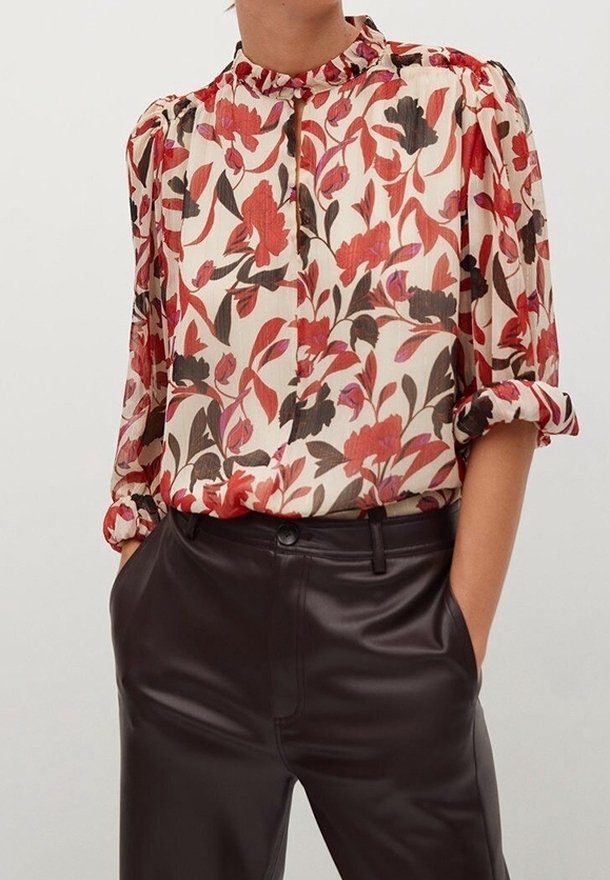 Personne portant une blouse à manches longues à motifs floraux rouges, roses et bruns, rentrée dans un pantalon en cuir marron foncé taille haute.