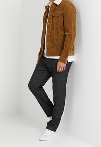 Brun fløjlsjakke med hvid fleecekrave, hvid t-shirt under, grå tapered bukser og hvide sneakers.