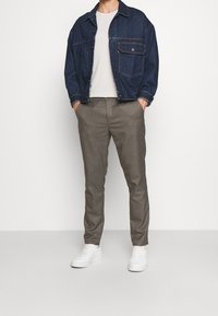 Mężczyzna w ciemnoniebieskiej dżinsowej kurtce, białej koszulce, dopasowanych spodniami w kratę i białych sneakersach, stoi z rękami w kieszeniach.