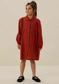 Rostfarbendes, geripptes Kleid mit Kragen, Knopfleiste und gerafftem Taillenbereich. Lange, gepuffte Ärmel und runder Saum. Schwarze Schuhe.