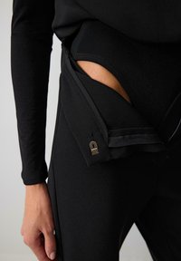 Melnas sporta bikses ar elegantu dizainu, kurām ir rāvējslēdzēja kabatas un gluda, elastīga tekstūra. Iekļauta neliela metāla detaļa.