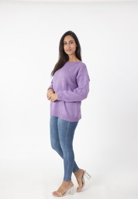 Lichtpaars sweater met verlaagde schouders en geribbelde zoom, gecombineerd met blauwe skinny jeans en doorzichtige hakken. Effen witte achtergrond.