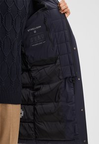 Person, der einen offenen navy-blauen Steppmantel hält, der das schwarze Innenfutter mit Strellson-Branding und einer Reißverschlusstasche innen zeigt.