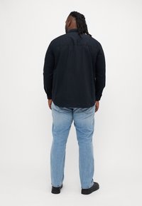Homme avec des dreadlocks portant une chemise noire à manches longues, un jean bleu clair et des chaussures noires, debout de dos sur un fond blanc.