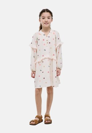 Jeune fille aux cheveux tressés portant une robe rose clair à manches longues avec imprimé floral et des sandales marron, debout et souriante.