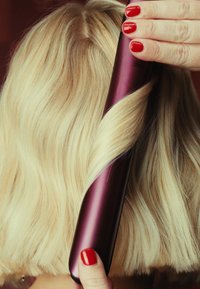 Hårlocktång med en slank, metallisk lila kropp och ett smidigt handtag, används för att skapa vågor i ljust blont hår. Röda naglar syns.