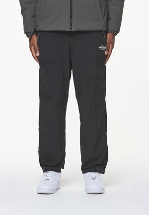 Person trägt schwarze Cargo-Hose mit Reißverschlusstaschen und weiße Turnschuhe, steht vor einem schlichten hellen Hintergrund.