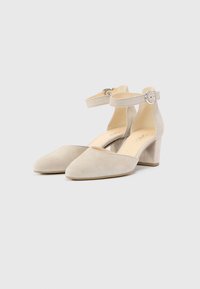 Chaussures beige en daim avec bride aux chevilles, bout pointu et talon bloc de hauteur moyenne. La bride est ornée d'une boucle en métal.