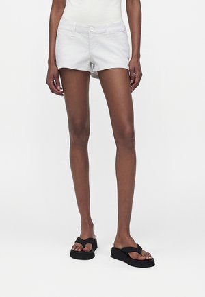 Fehér rövidnadrágot és fekete platform papucsot viselő személy áll egy egyszerű, fehér háttér előtt.